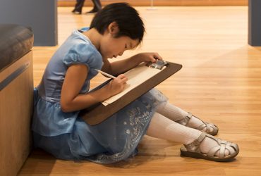 A young visitor wearing a blue dress sits on the floor to sketch in a gallery at the Nasher Museum.