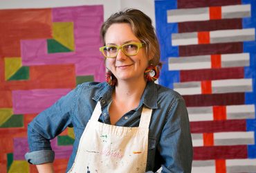 Artist Martha Clippinger poses with right hand on hip, wearing yellow-rimmed glasses, in front of one of her colorful textile works.