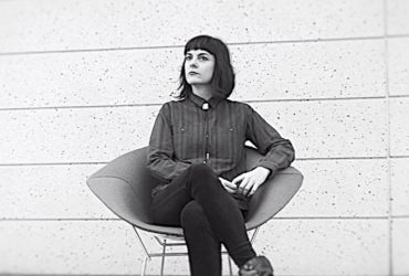 Black-and-white photo of Casey Cook sitting in a chair, a still from the video by J Caldwell.