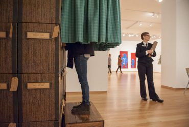 A visitors' legs peek out from under the green curtain of Stacey L. Kirby's Power of the Ballot. The artist stands nearby with a clipboard.