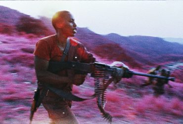In this film still, a male figure points a rifle to the right. The landscape is pink and purple because of infrared film.