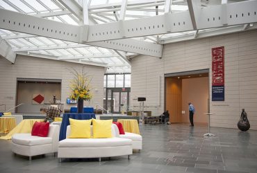 The Nasher Museum Great Hall is set up for the opening of Open This End. A security guard stands at the entrance to the pavilion. White couches have red and yellow pillows.