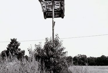 Black-and-white photo of a car high up on a pole.