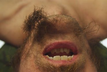 George Jenne, Knowing Me (knowing you)(still), featuring a man's upside-down half-bearded face, mouth open, teeth exposed.