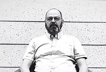 Black-and-white photo of Bill Thelen, seated in a chair, a still from the video by J Caldwell