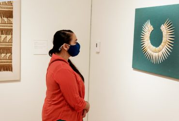 A Duke Student stands in the galleries wearing a protective mask during COVID-19. She looks at a large shell collar of the Nazca people's of Peru; this work is part of the Nasher Museum's Mesoamerican collection.