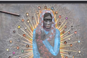Mixed-media portrait of a seated figure in a blue garment and patterned head covering, surrounded by radiating lines and small photo portraits on a gray background.
